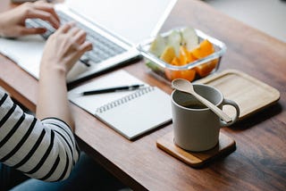 A woman is typing on her laptop. A notebook, cup of coffee, and fruit sit closely on her desk. #freelancing #writing #workfromhome #writer #wfh