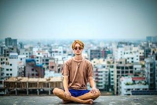 Young man in sunglasses sitting cross-legged on the ground against a background of buildings