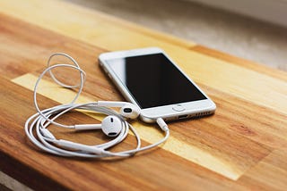 Earbuds are connected to a cell phone through the usb port — earbuds and phone rest toward the edge of a wooden desk