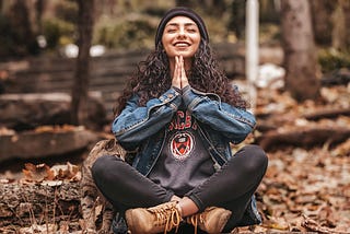 A woman with long dark hair sitting cross-legged with hands in a praise symbol smiling