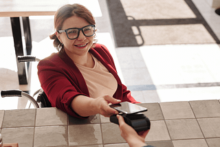 A smiling woman using a wheelchair holds her smartphone over a payment device at a cafe.