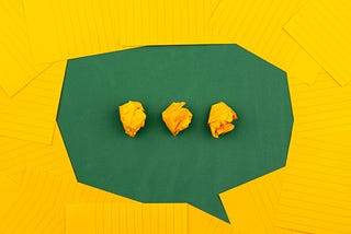 An artistic depiction of a speech bubble with three dots inside the bubble. The image was made with sheets of yellow ruled paper against a green background.