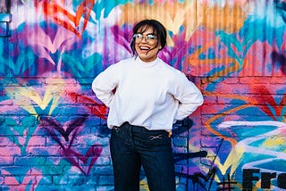 a woman wearing specs, white top and black jeans infront of a really colorful background.