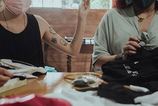 foto em close up de duas mulheres, sem mostrar o rosto. Estão bordando em uma mesa de madeira ehá tecidos e ferramentas sobre a mesa.