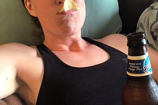 A woman laying on a couch with a chip in her mouth, holding a beer.