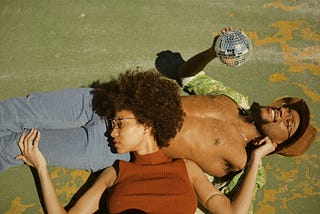 Young man lying on his back in the sun looking at a small glitter ball in his hand. Young woman lying on her back, across him, her head on his stomach