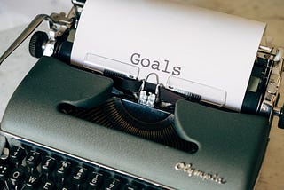 An antique typewriter on a wooden desk with a sheet of paper in it with “Goals” typed on it.