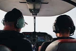 Pilot and co-pilot sitting in the cockpit with headphones on viewed from the back of their heads and out the cockpit window.