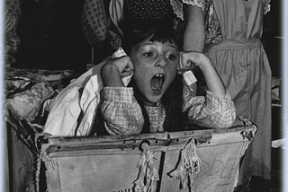 Black and white photo of a community theatre production of Annie. Molly in a laundry basket, singing. Three orphans stand singing behind her.