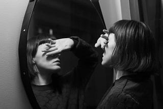 A black & white image of a lady looking into a round hanging mirror covering her eyes to stop her from seeing her reflection