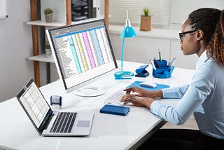 Professional Black woman sitting at a desk, looking at computer screen with a detailed spreadsheet.