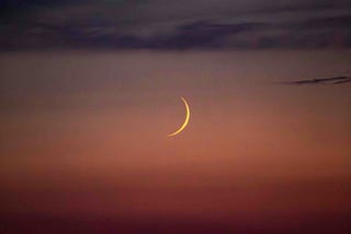 A crescent moon is visible in the evening sky, surrounded by a gradient of deep orange and purple hues. The sky transitions smoothly from the remnants of sunset at the horizon to the darkening night above. The scene is tranquil, with a sense of calmness as the moon's slender arc stands out against the dimming light, creating a serene and peaceful atmosphere.