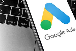 Google Ads logo in blue, yellow, and green on a smartphone, with a partial laptop keyboard featuring black and silver keys.