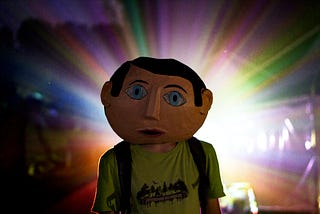 Rainbow of colors comes out from behind a man in a t-shirt and vest, wearing a large mask of a face with soulful eyes and a worried, curious expression.