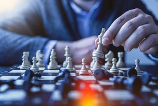 A faded upper chest of a man in a blue suit jacket and light blue shirt holding a white chess piece between two fingers over a full dark blue and white chess board of pieces. Writer Christine McDonald uses the picture to symbolize the strategy it takes to win.