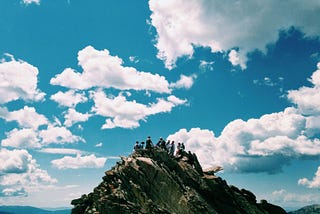 Rocky mountain top with a bunch of people visible against the skyline. Blue sky. Scatered white clouds.