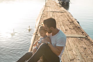 A father holds his daughter on a dock at the lake with the sun glistening behind them