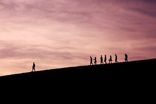 Silhouette of people walking down a hill with one person out in front of the group.