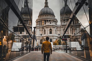 Image; man walking along a city street, wearing a short jacket and jeans, facing a church tower with his back to the viewer