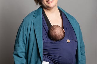 Stella facing camera and smiling with young baby in sling.