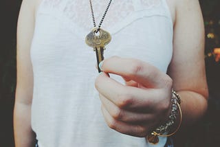 a woman in a white blouse holds a key that’s on a chain around her neck. we only see her torso. not her head.