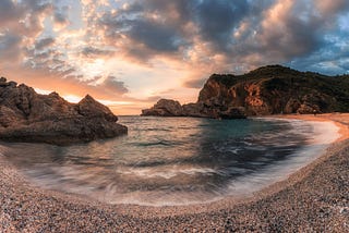 A landscape photo of some water moving between two irregular shaped rocks, there is a pink-orange and blue tinge to the rocks, water and sky that makes one think of serenity.