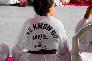 A student of TaeKwonDo, wearing a white uniform, sits on a mat with others looking towards their right.