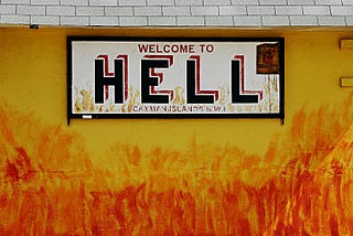 A sign reading “Welcome To Hell, Cayman Islands” hanging on a wall coloured like it is set aflame.