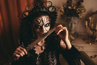 A woman dressed in black with eyes outlined in black holds a knife to her lips
