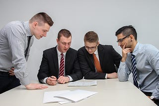 Four men wearing suits and ties sitting at a table reviewing a contract or some form of paper.