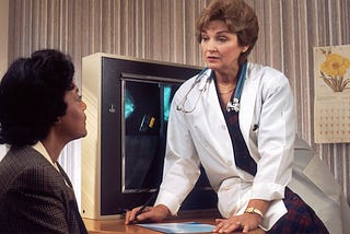 A middle aged white woman wearing a white coat with a stethoscope is leaning over a table in front of a computer showing an XRay while talking to a black woman sitting on the other side of the table.