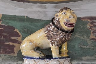 A ceramic lion on a marble mantle shelf. The lion is naively made with an almost human face, and glazed in with yellow body and face and brown mane. The glaze is chipped in places. Around its neck is a circle of small back beads. Behind the lion, the wall is roughly but richly painted in earthy green, brown, yellow, grey and white.