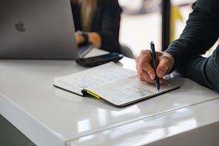 Person writing notes on a notepad on a desk next to a laptop