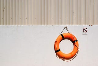 A bright orange lifebuoy hanging from a rope against a cream colored wall.