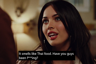 Jennifer (Megan Fox) in white jacket and hoop earrings looking up at Chip (offscreen). The caption (with a black background and white text) underneath says “It smells like Thai food in here. Have you guys been f***ing?”