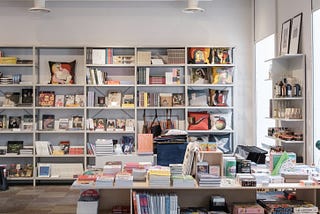 A brightly lit bookstore, with white shelves and a white table, both full of books for sale.