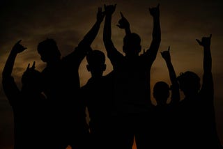 A photo of six boys or young men with their arms raised up. They are silhouetted so they each appear very dark and their facial features can’t be seen. Each is making a sign with their fingers, apparently a victorious sign.