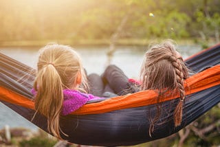 Two girls sharing a hammock on a summer day spent mostly outside. The one on the left is gazing at a lake and we seeherlong unruly ponytail in light brown hair. The second girl a cousin of almost the same age has long hair as well but a top section has been pulled back gathered with an elastic and braided. The sun glows from the top left.