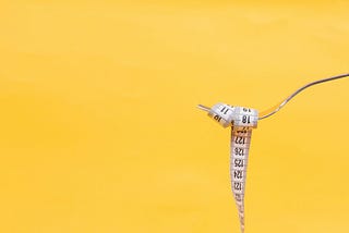 A fork in front of a yellow background, holding a tape measurer that is wrapped around the tines like a piece of pasta.