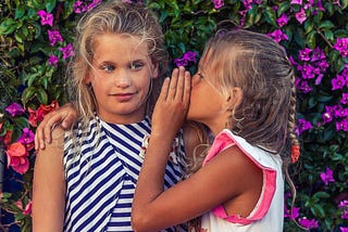 Two young girls with one whispering something, possibly gossip she’s heard. in the ear of the other.