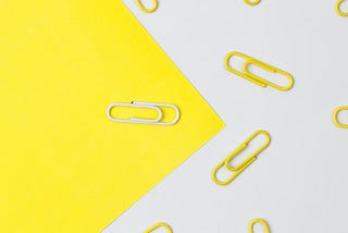 Piece of yellow paper on an angle with one white paperclip on it against a white background with many yellow paperclips on it