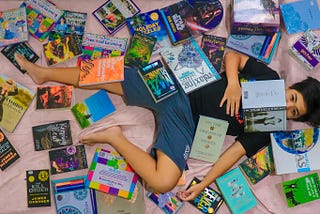 a woman lies on a bed with books all around her and on top of her. one open at eye level she’s engrossed in.