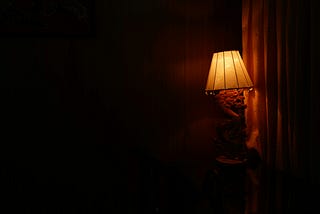 A darkened bedroom illuminated only by a low-powered bedside lamp, casting a soft, warm glow in the otherwise shadowed room