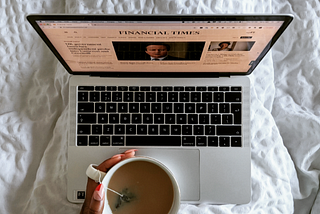 Laptop showing the Financial Times homepage on a bed with a cup of tea in hand and funky red and pink nails