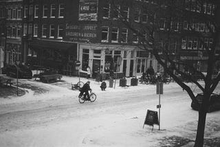 A cyclist going along a winter urban street, with the street covered in snow. The photo is black and white and taken from far away; in the foreground are trees on the side of the street, and behind the cyclist is an intersection and a row of three-story retail stores