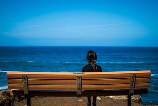 Woman with black hair sitting on a wooden bench in front of the ocean.