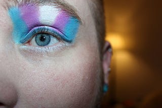 A close up of a human eye, with transgender flag colours applied as eye shadow.