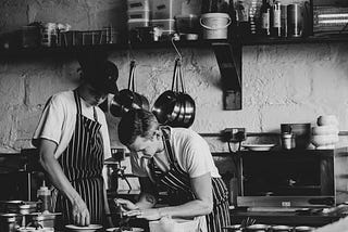 Two men, workers in a restaurant, with aprons on are in a kitchen preparing food.