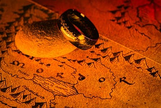 A glowing gold ring with Elvish inscriptions rests on a rock, placed over a map of Mordor from “The Lord of the Rings,” illuminated by fiery orange and red light.