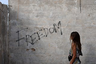 Lightly black spray-painted word “FREEDOM” on a concrete wall being stared at contemplatively by a young women whose face is hidden by her long hair giving the sense more of her staring at the meaning of this word.